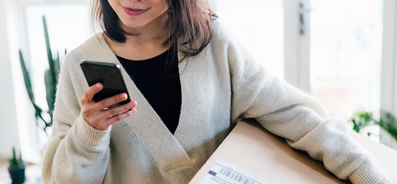 A woman holding a big brown box/package in one hand and looking at her smartphone that's in the other hand