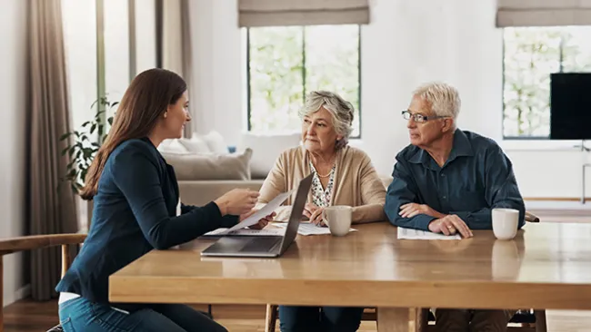 A young woman sitting at a laptop talking to an elderly couple at a kitchen table.