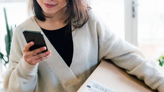 A woman holding a big brown box/package in one hand and looking at her smartphone that's in the other hand