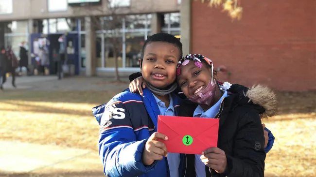 Two children holding a letter in front of a school