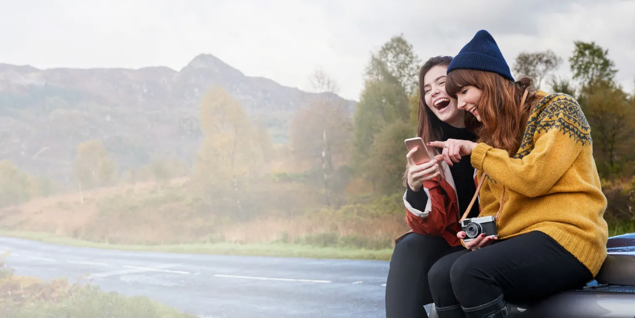 Two woman sitting by a river laughing at what's on their phone screen