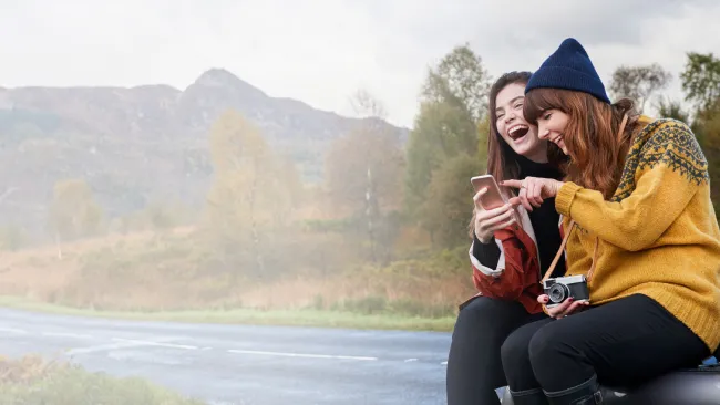 Two woman sitting by a river laughing at what's on their phone screen