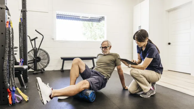 Man on the floor using a foam roller to stretch his legs as a physical therapist kneels beside him.
